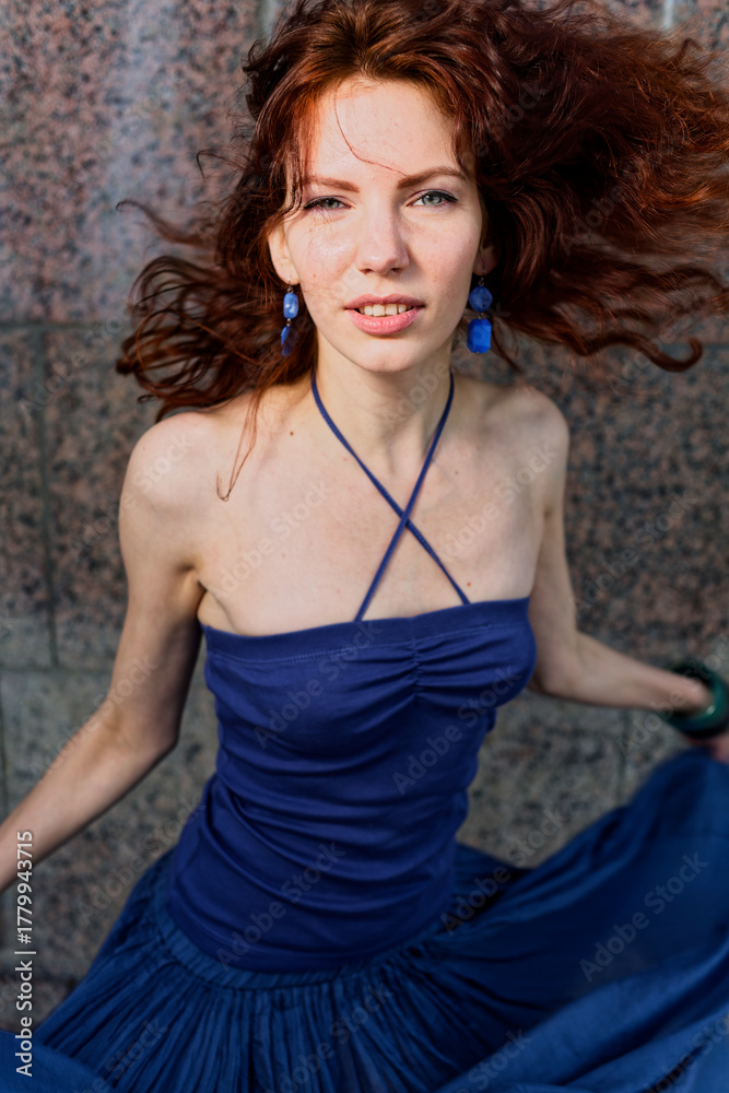 Fototapeta premium Confident Redhead Posing in Vibrant Blue Dress Against Industrial Urban Wall, Windblown Hair, Chic Fashion Shot