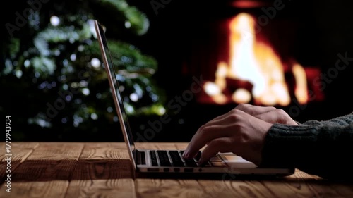 A man works on a laptop in front of a fireplace and a Christmas tree. Freelancing, remote work, or studying at home during the holidays. A warm festive atmosphere for New Year or Christmas.