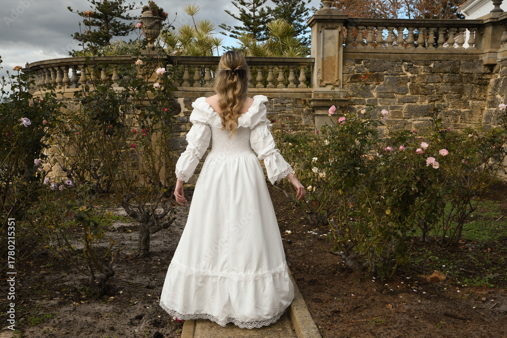 Naklejka premium portrait of beautiful young blonde model wearing historical white wedding gown. ethereal back view figure, walking away in romantic rose garden of fairytale castle garden background