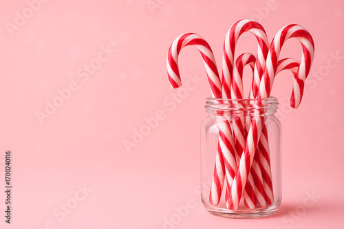 A vibrant and sweet Christmas scene. On the right side, a clear glass jar filled with colorful red and white candy canes. The left two-thirds of the image is a clean, minimalist, soft pink background.