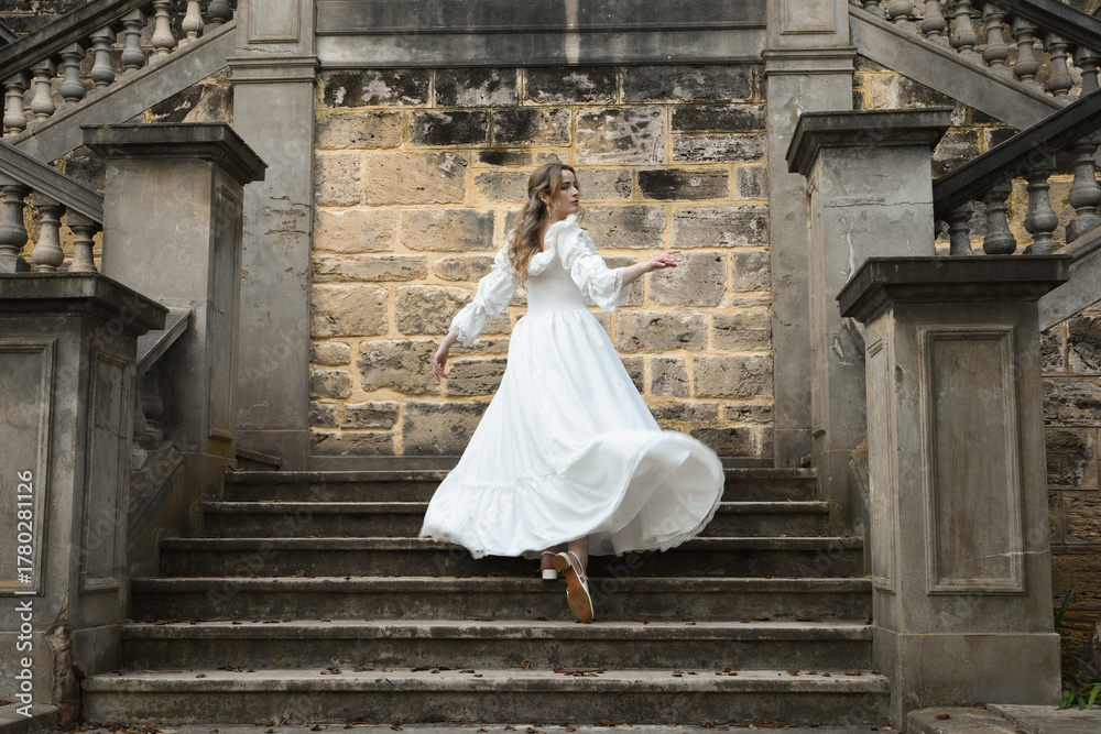Naklejka premium Full length female figure of blonde model wearing white fantasy wedding gown. Back view pose, walking away on historical staircase of castle location 