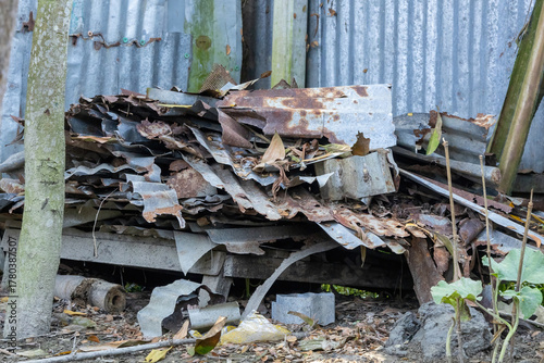 Weathered and deeply rusted corrugated metal scraps, bent and broken, are stacked in a disorganized pile next to a house in a poor village setting in Bangladesh. Abandoned junk and texture.
