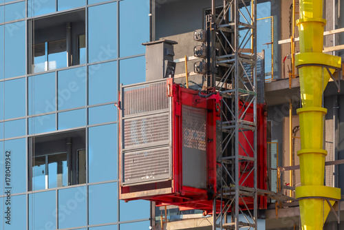A red construction elevator rising along the facade of a modern building and a yellow plastic pipe for dumping construction trash