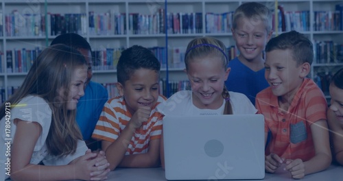 Smiling schoolchildren gathering around laptop in library, with bookshelves backdrop