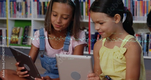 Reading two schoolgirls crosslegged using tablets on library carpet, with bookshelves backdrop