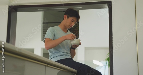 Eating teenage Korean boy scooping food from white bowl on kitchen counter ledge, with potted plant