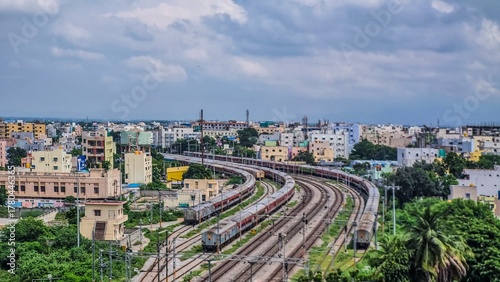 Curved railway tracks lead into the heart of Tirupati city, surrounded by colorful residential blocks and lush greenery. The cloudy sky adds a calm monsoon mood to this vibrant and fast-growing city