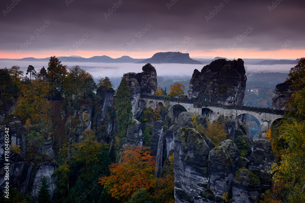 Fototapeta premium Bastei bridge, Sächsische Schweiz, Germany autumn landscape. Travel in Europe. Misty foggy morning sunride in nature. Rock hills with trees and forest. Elbe Sandstone Mountains, fog mornig with clouds