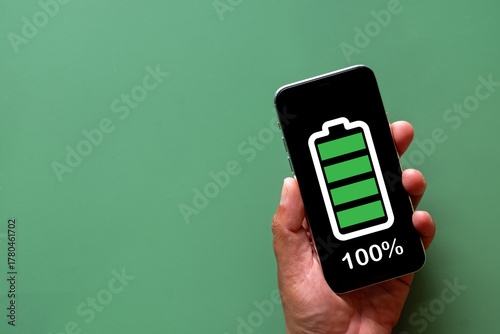 Hand holding a smartphone with a 100% fully charged battery icon on the screen. Concept of full power, energy, and readiness. Top view on green background with copy space.