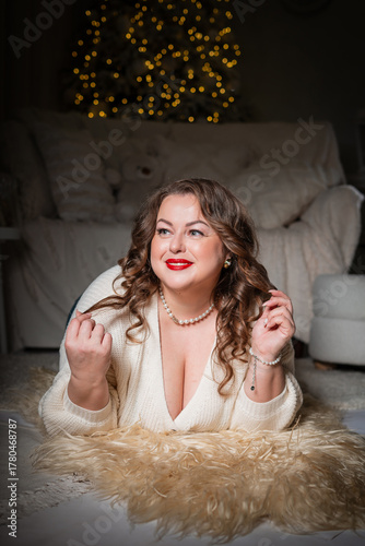 A portrait of a beautiful,chubby  woman with a sweet smile against a New Year's backdrop. A buxom woman in a white knitted sweater with an open neckline poses against a backdrop of Christmas lights.