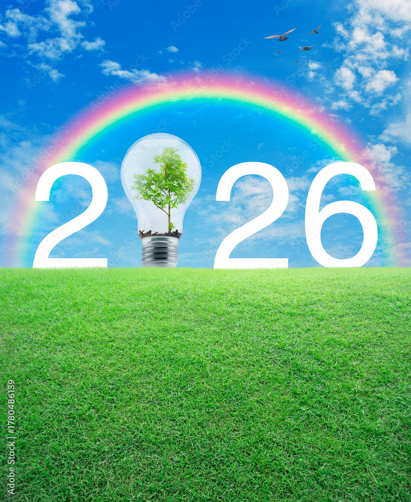 Naklejka premium 2026 white text and light bulb with tree inside on green grass field over blue sky, white clouds and rainbow, Happy new year 2026 ecological cover concept