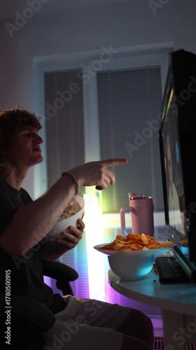 Vertical screen student eating popcorn and chips while watching videos on a glowing monitor at home showing relaxed evening mood casual lifestyle and little focus on healthy habits or study