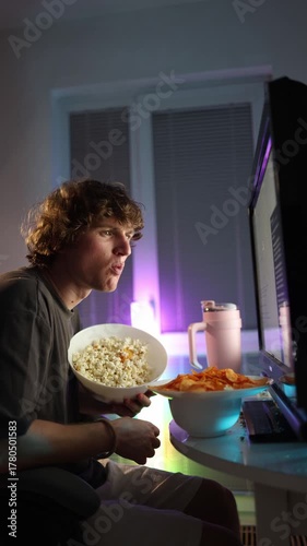 Vertical screen young man eating popcorn and chips while watching a glowing monitor at home creating a relaxed evening vibe casual lifestyle moment of leisure and digital entertainment