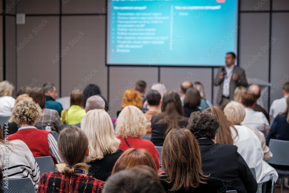 Obraz premium Audience professionals attend live presentation in bright conference room. Focused attendees watch speaker share insights while slides illuminate background