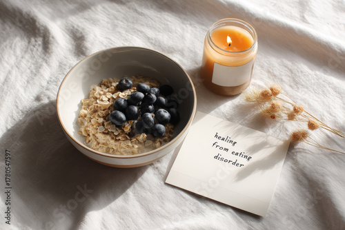 A bowl of oatmeal with blueberries next to a lit candle and a note reading healing from eating disorder on a white wrinkled sheet symbolizing recovery, self-care, and mental health