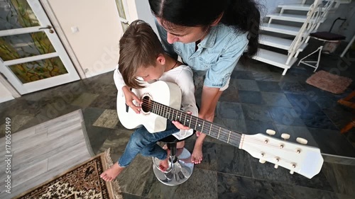 A young boy sits on a stool, focused on strumming a white guitar under the guidance of a caring adult, surrounded by a stylish home setting.