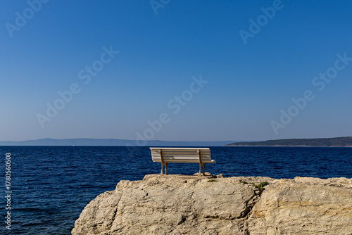 Fototapeta Naklejka Na Ścianę i Meble -  A bench overlooking the sea on a rocky shore near a walking path. Adriatic Sea, Makarska, Croatia.