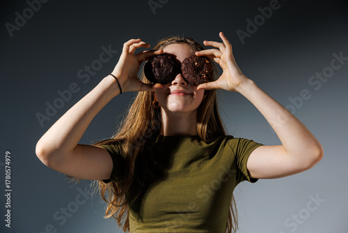Cheerful girl playfully holds two cookies over her eyes like glasses, smiling against a gray background. A fun and light-hearted food-themed portrait