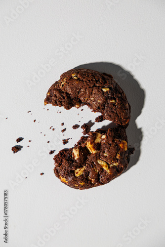 A dark chocolate cookie with visible nut chunks is split in half, with crumbs scattered across a white background, creating a dynamic and appetizing composition