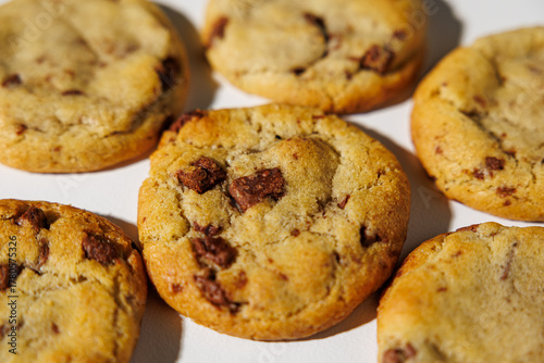 Photos A detailed view of golden brown chocolate chip cookies arranged on a white surface, showing melty chips and soft texture under natural light