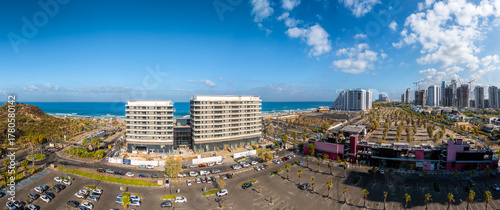 Aerial view of the coastline meeting modern buildings, a large car park, and high-rise buildings under a blue sky, Rishon LeZion, Center District, Israel.