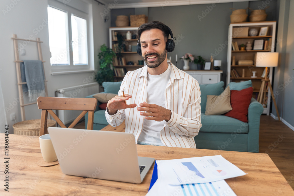 Obraz premium Young freelancer leading an online video meeting from a cozy home office smiling man with headphones gestures while speaking on a laptop during remote consulting and sharing project results.