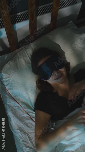 Vertical video. Woman lying in bed preparing for healthy deep sleep by inserting earplugs in her ears and wearing a sleep mask