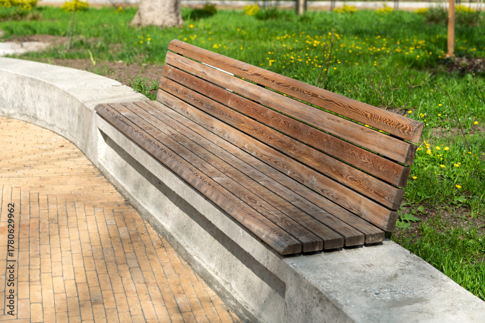 Obraz premium Curved park bench with wooden slats and concrete base sits empty on a sunny day in a blooming green urban garden.