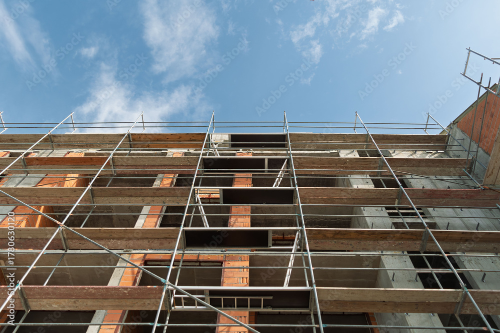 Fototapeta premium Low angle scaffolding grid on unfinished concrete building with orange brick columns visible through openings under blue sky.