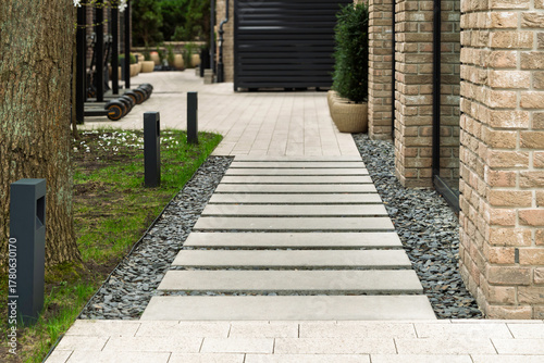 Modern walkway made of concrete slabs leads along landscaped area with decorative gravel and brick building, featuring outdoor lighting.