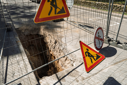Deep trench along city street surrounded by safety fence and warning signs, concept of road construction, repair and infrastructure safety.