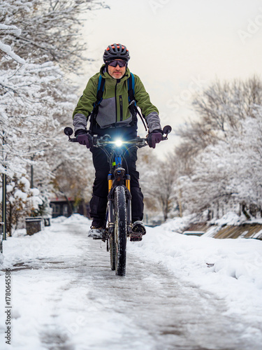 A man rides a bicycle on a winter day along a sidewalk surrounded by snow-covered trees. The man is wearing a bicycle helmet. An active lifestyle in winter.