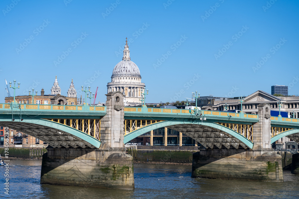 Fototapeta premium Southwalk Bridge stands tall over the River Thames on a clear sunny day. The iconic structure connects the banks of London, showcasing its unique architecture and lively surroundings.