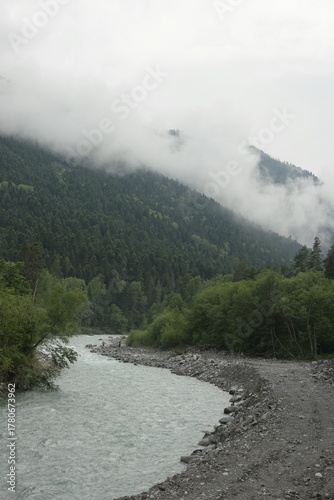 A stormy mountain river flows between rocks, boulders and cliffs in Arkhyz against a backdrop of mountains and dense forests, hidden by clouds and thick fog.