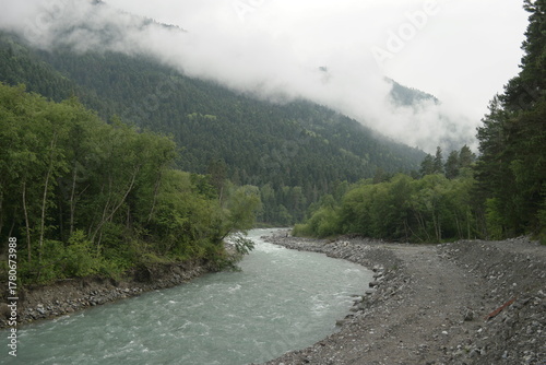 A stormy mountain river flows between rocks, boulders and cliffs in Arkhyz against a backdrop of mountains and dense forests, hidden by clouds and thick fog.