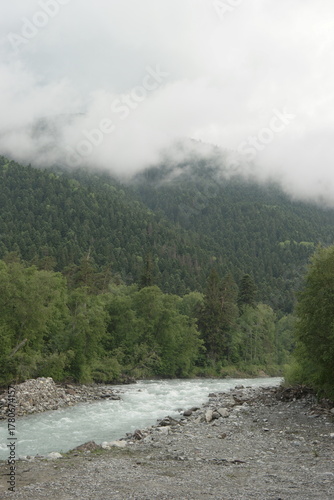 A stormy mountain river flows between rocks, boulders and cliffs in Arkhyz against a backdrop of mountains and dense forests, hidden by clouds and thick fog.