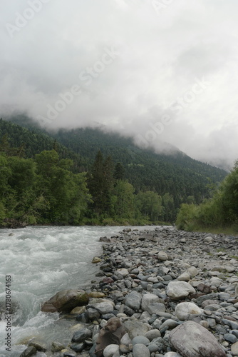 A stormy mountain river flows between rocks, boulders and cliffs in Arkhyz against a backdrop of mountains and dense forests, hidden by clouds and thick fog.