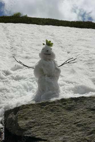 A lone snowman with arms made of twigs, eyes made of stones, hair made of grass leaves, and a mouth made of a stick stands on a mountain against a backdrop of snow and rocks, stones