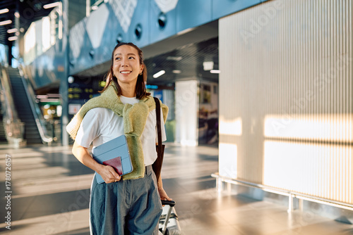 Smiling woman with passport and tablet pulling luggage, arriving or departing from modern airport terminal