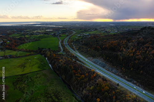 Aerial view of the M1 motorway with cars surrounded by autumn forest and green fields during a cloudy evening in rural Ireland