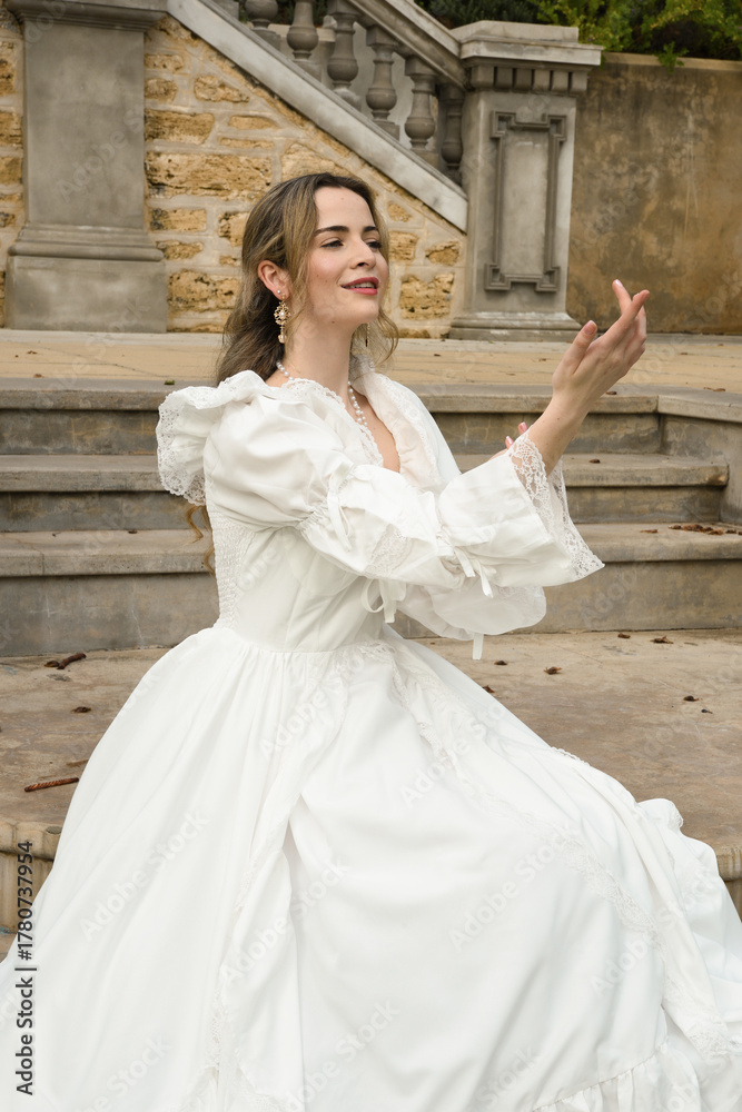 Obraz premium figure of female blonde model wearing white fantasy wedding gown. romantic sitting pose on staircase of historical fairytale castle location