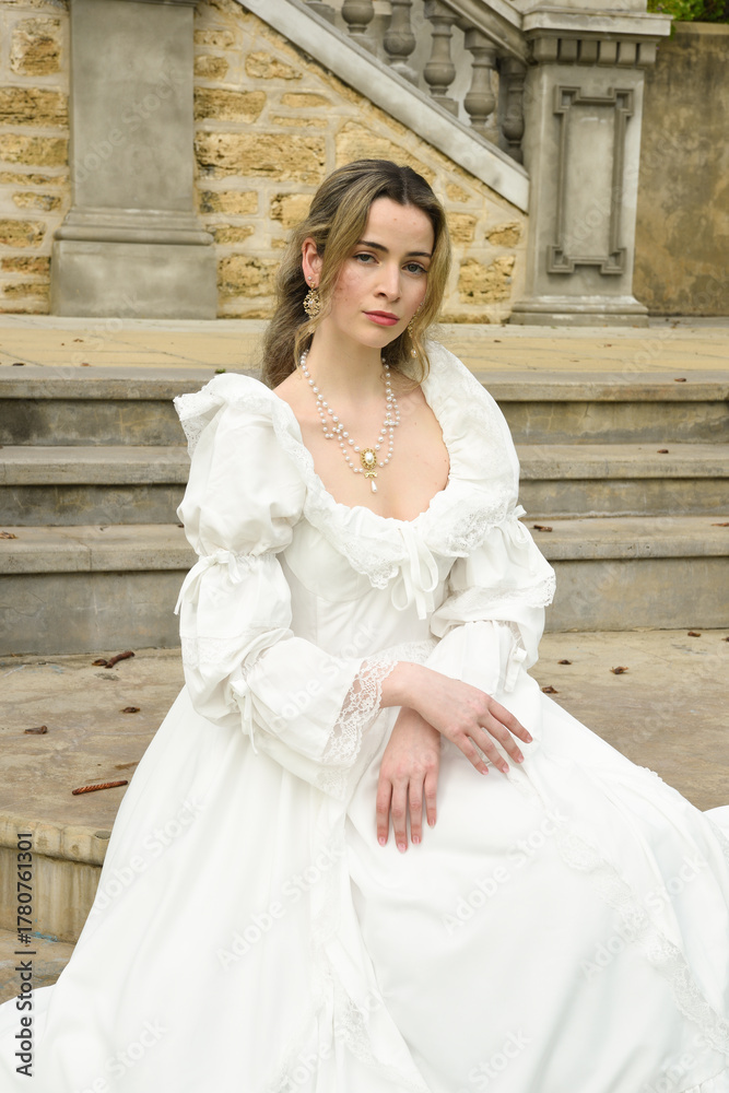Obraz premium figure of female blonde model wearing white fantasy wedding gown. romantic sitting pose on staircase of historical fairytale castle location 