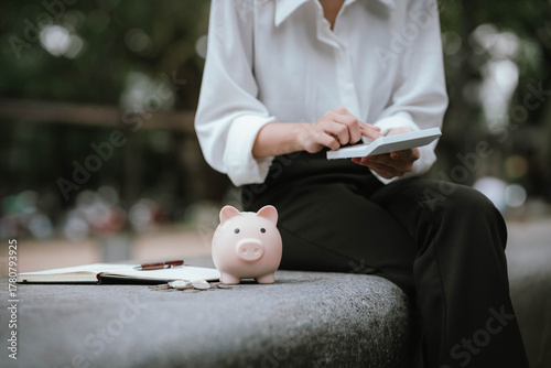 A person saves money by putting coins into a pink piggy bank while holding a calculator, symbolizing financial planning, saving, and budgeting.