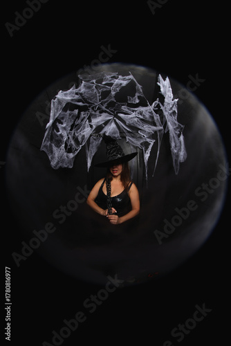 portrait of a young witch in hat on black background with spider net and holding chain  in her hands