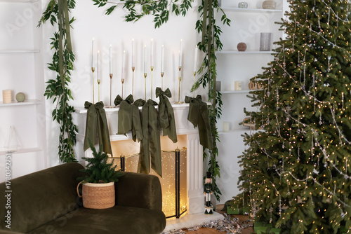 Festive decorated white fireplace with candles with ribbon green bows in interior modern living room. Xmas at home. New year decor. Mantelpiece with garland witn branches	
