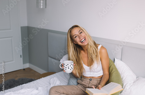 Fotomural Cheerful woman laughing while reading a book and holding coffee mug, enjoying an