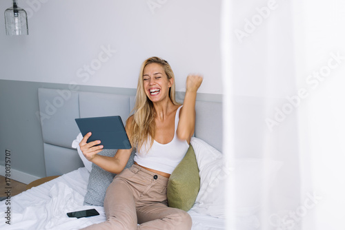 Fotomural Cheerful woman celebrating a digital success while sitting in bed with tablet, e
