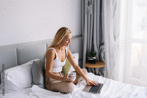 Fotografía Smiling young woman typing on laptop while seated comfortably in bed, embodying
