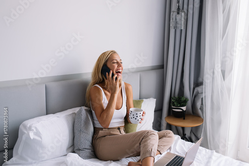 Fotomural Woman smiling during phone call while holding a mug and seated with laptop, high
