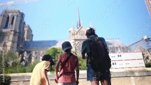 Family enjoying a summer trip to paris, standing by the seine river and observing notre dame cathedral undergoing restoration with construction cranes visible, symbolizing resilience and heritage